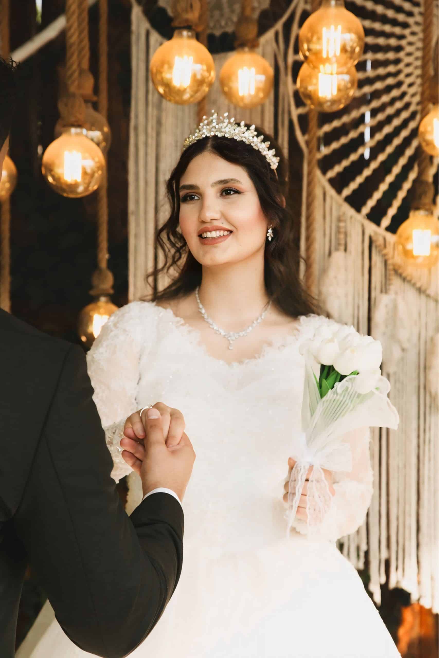 Une mariée souriante, en robe blanche, tiare et collier, tient un bouquet de tulipes blanches et la main d'un homme. Elle se tient sous des ampoules lumineuses et des décorations macramé.