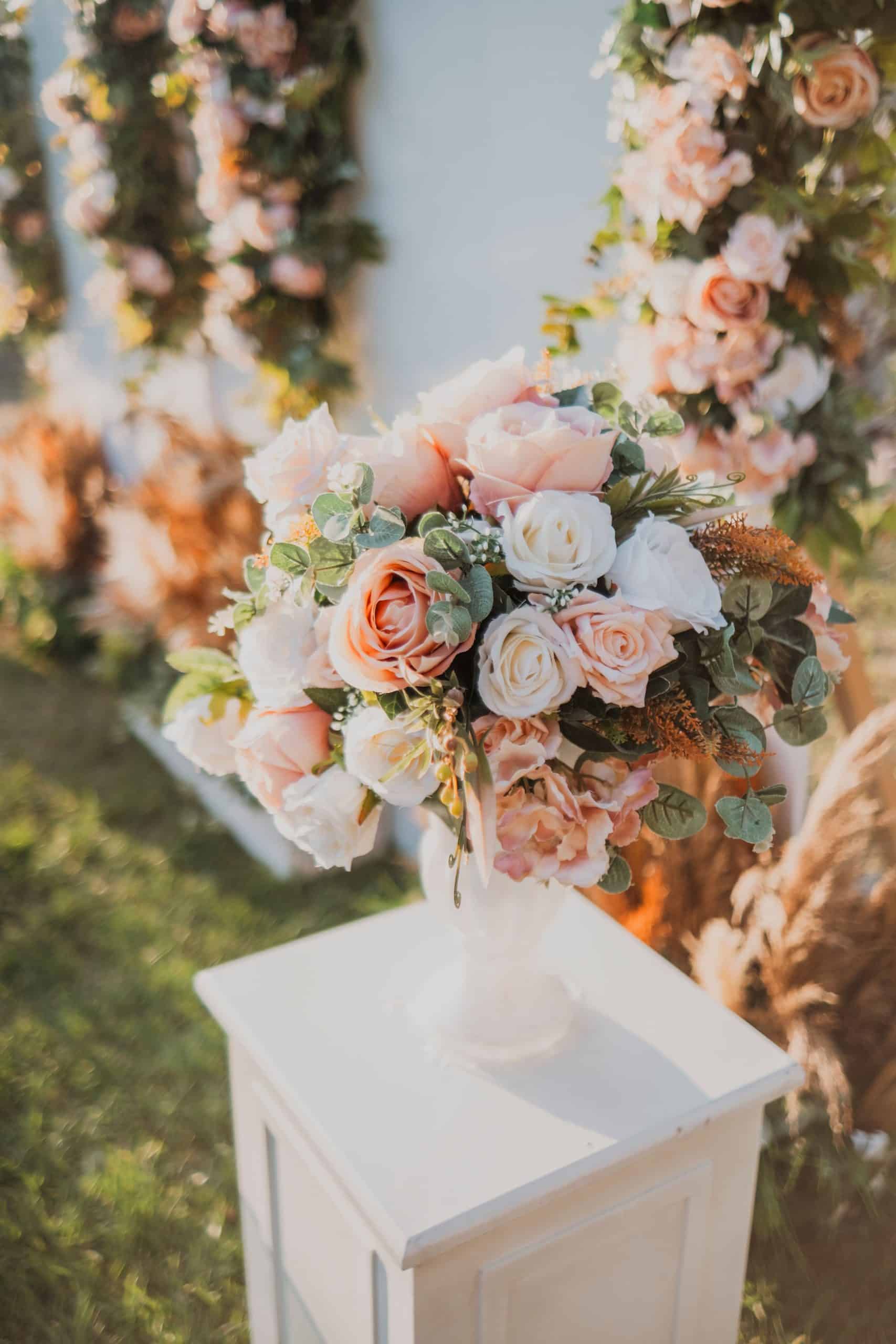 Un bouquet de roses pâles et blanches, avec feuillage vert, est posé dans un vase blanc sur un piédestal, entouré de décorations florales floues en extérieur.