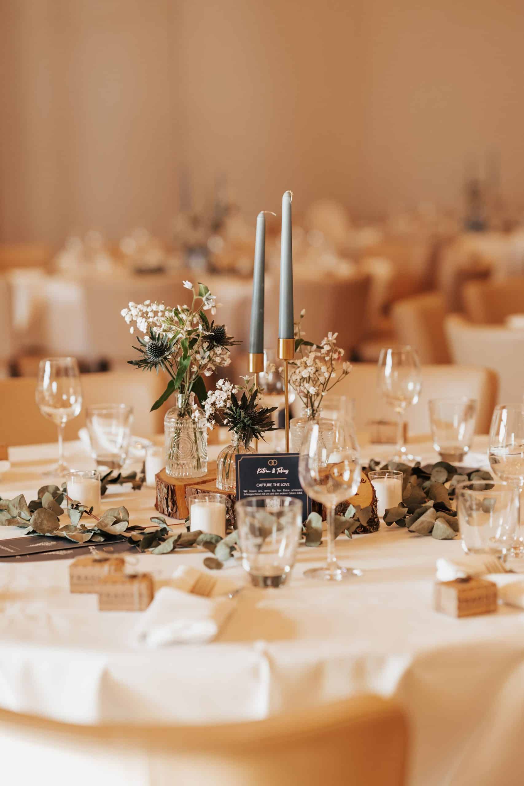 Centre de table de mariage avec bougies grises, petits vases de fleurs blanches et chardons, feuilles d'eucalyptus et bougies chauffe-plat, sur une nappe blanche. Texte : Katrin & Tobias, CAPTURE THE LOVE, Wir freuen uns, wenn du hier deine schönsten Schnappschüsse mit uns und den anderen Gästen teilst.