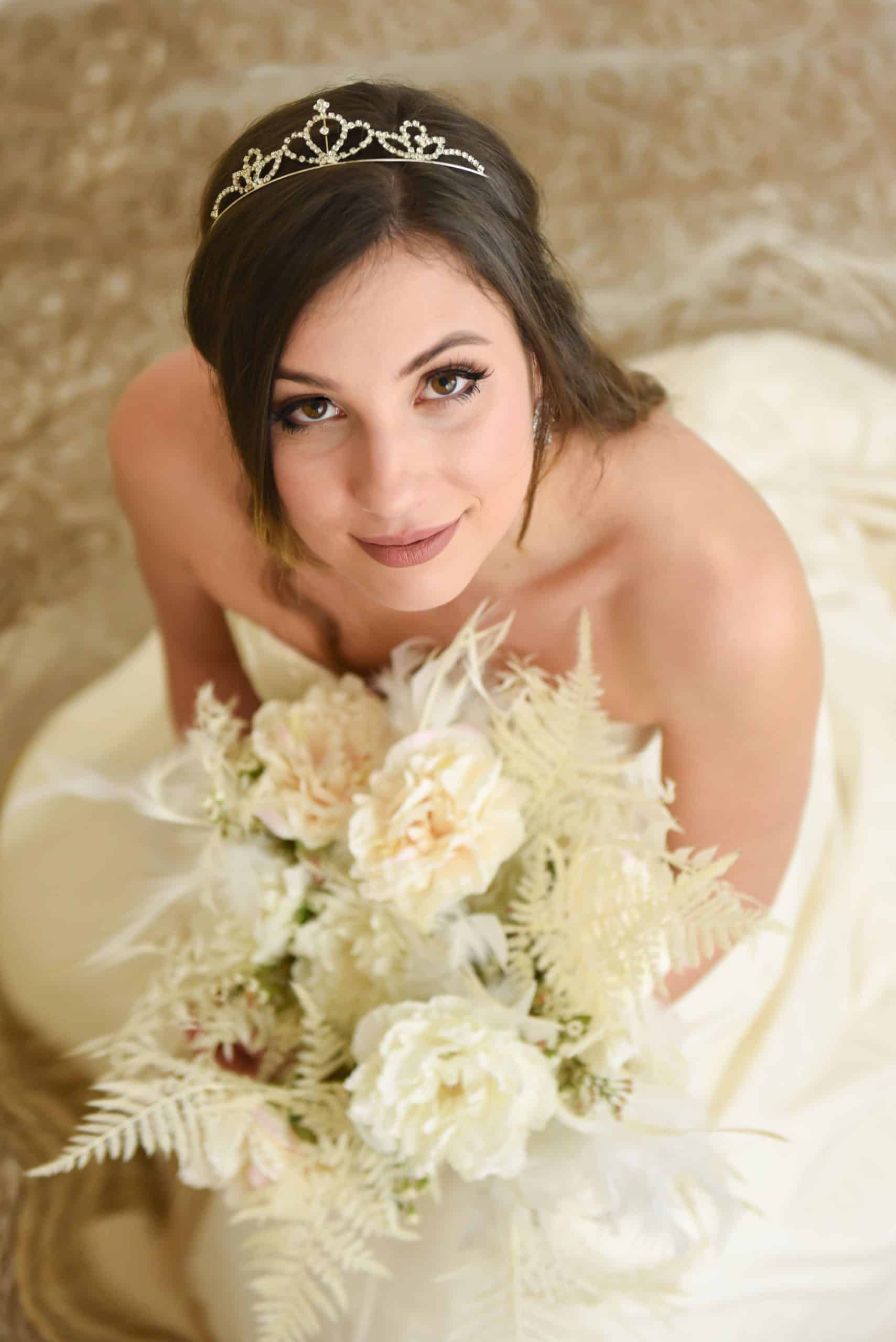 Une jeune femme en robe de mariée sans bretelles et diadème regarde vers le haut, tenant un bouquet de fleurs pâles et de feuillage. L'arrière-plan est clair et texturé.