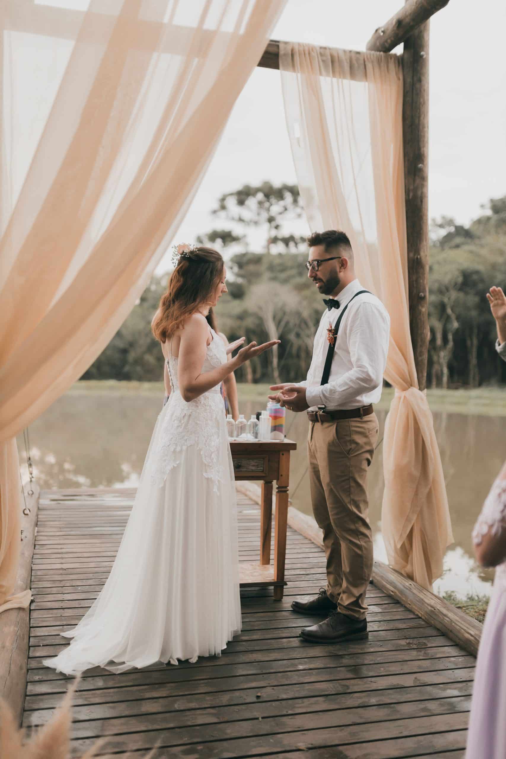 Couple en tenue de mariage effectuant une cérémonie de sable, se faisant face sur une estrade en bois. Des voilages beiges encadrent la scène devant un plan d'eau et des arbres.