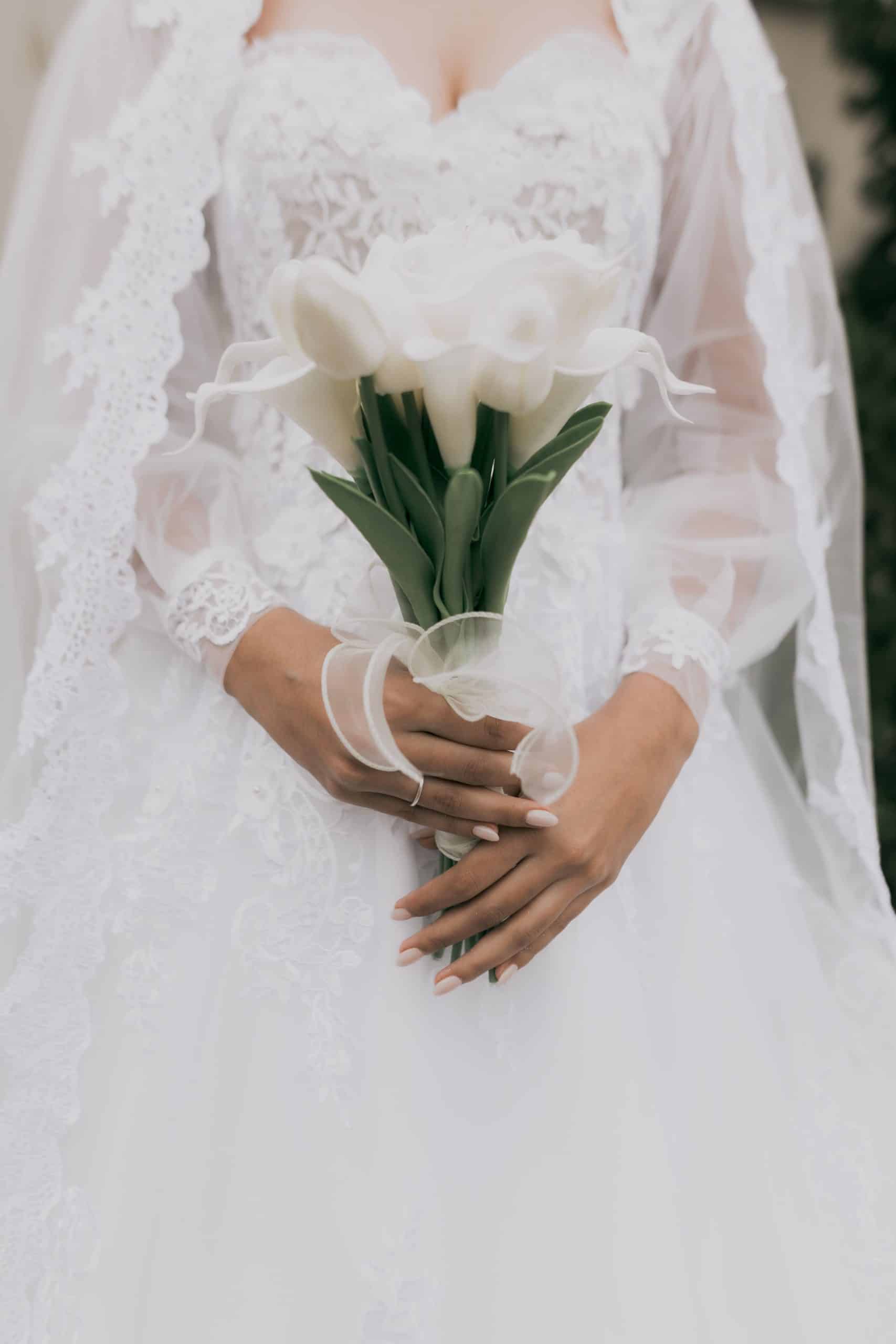 Une personne vêtue d'une robe de mariée blanche en dentelle et d'un voile transparent tient un bouquet de lys calla blancs, attaché avec un ruban.