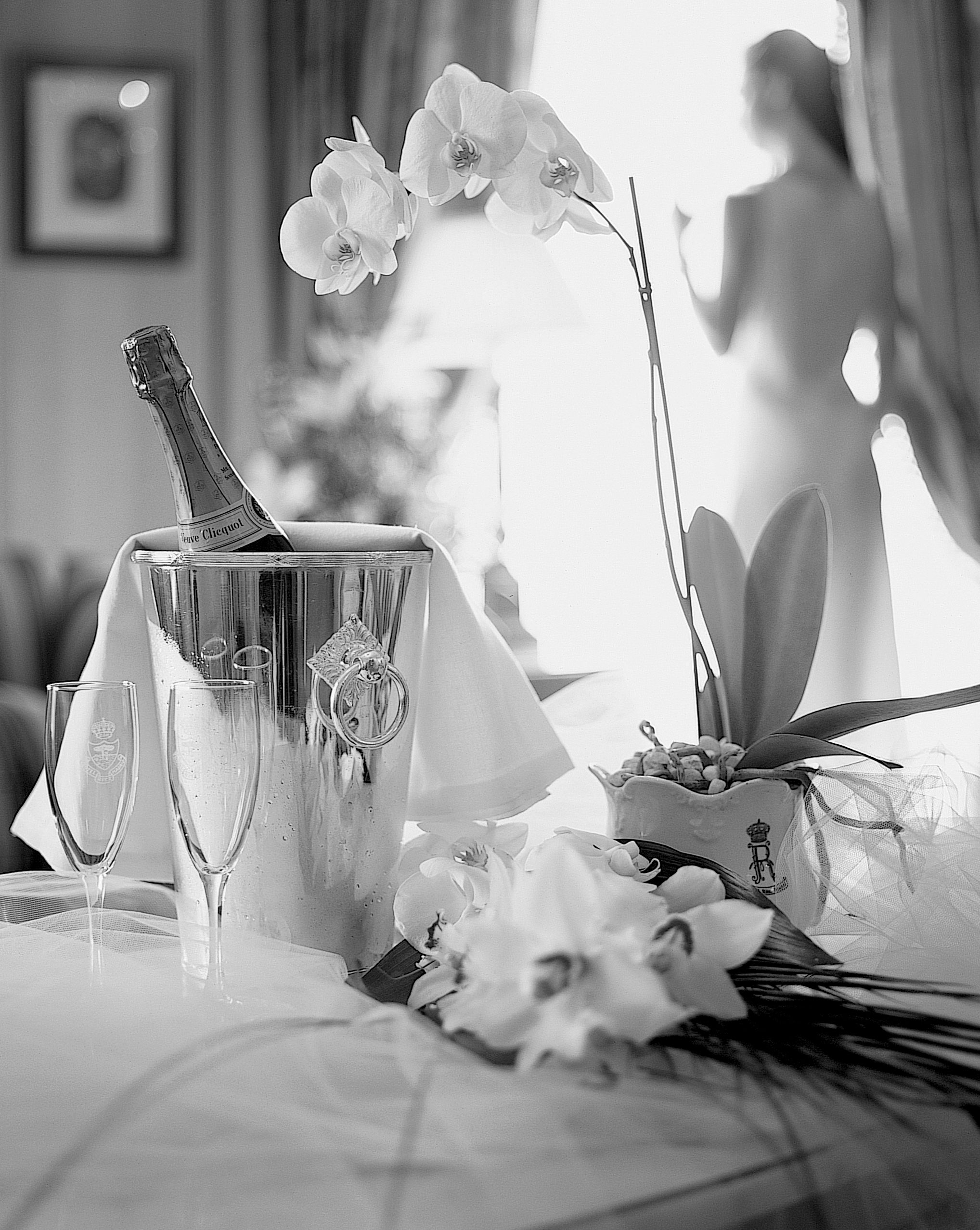 Bouteille de Veuve Clicquot en seau à glace, deux flûtes et orchidées sur table. Une silhouette floue se tient devant une fenêtre lumineuse. Pot: R ROYAL.