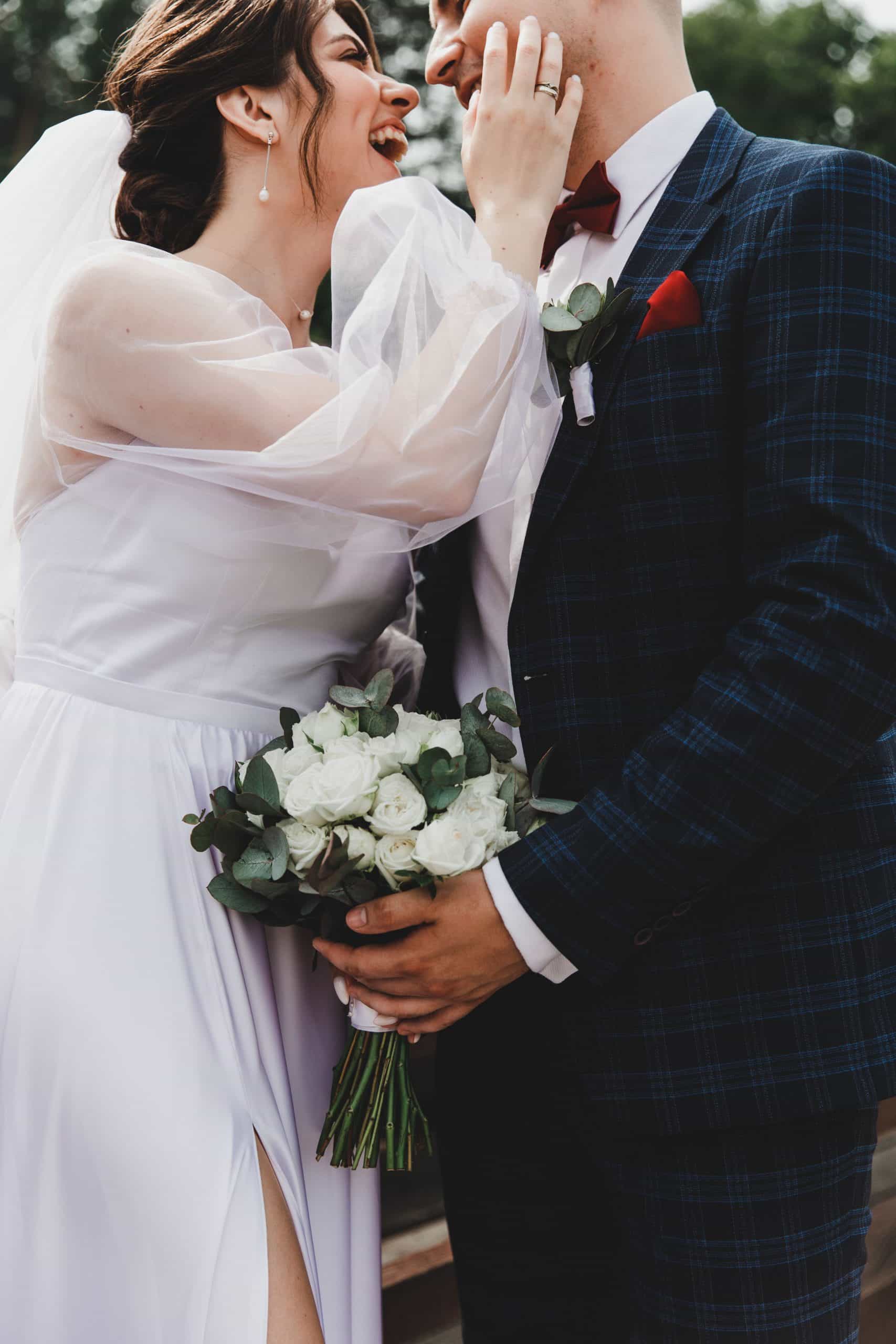 Une mariée rieuse en robe blanche, aux manches transparentes, touche la joue du marié en costume à carreaux bleu et nœud papillon, qui tient un bouquet de roses blanches et eucalyptus, en extérieur.