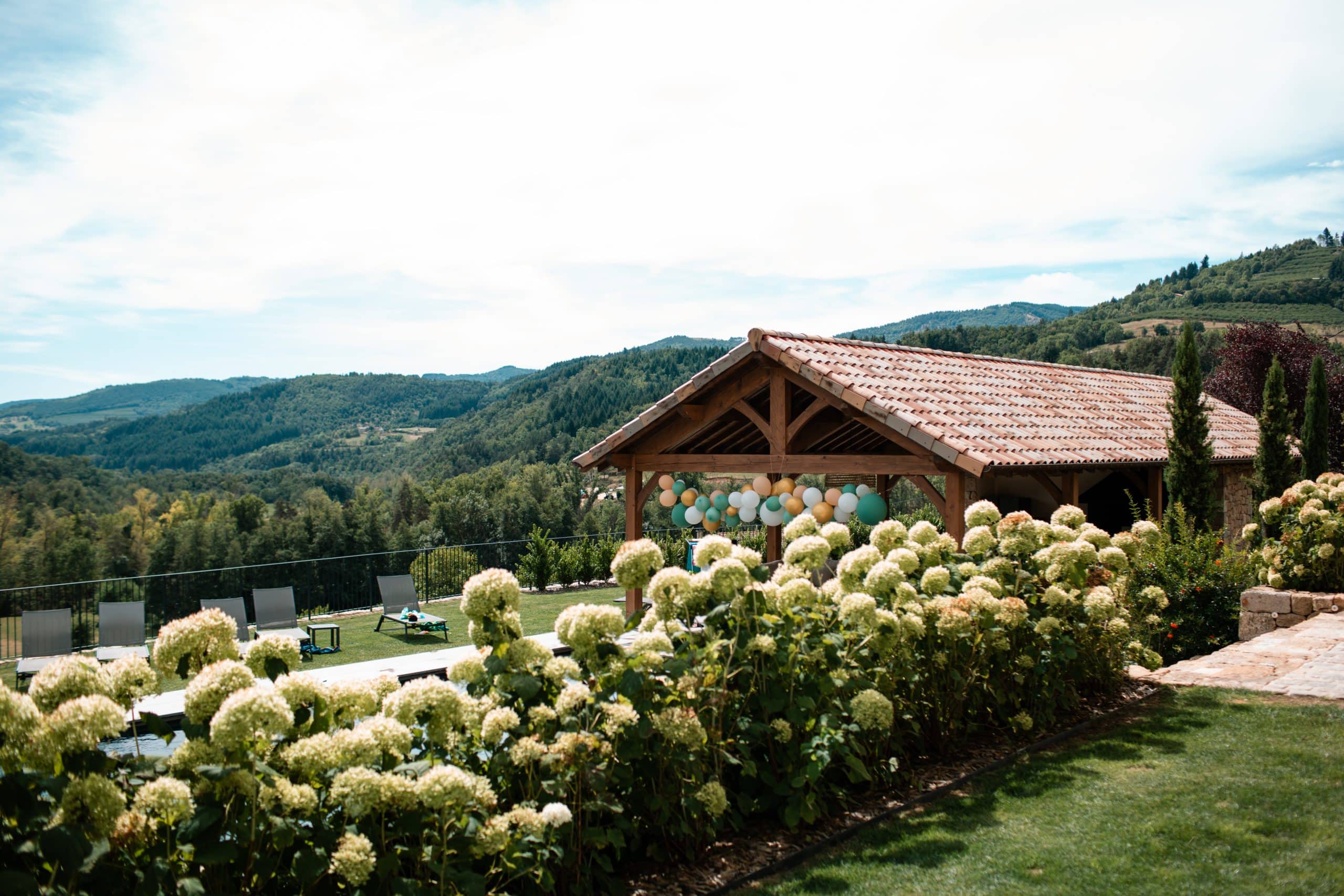 Un gazebo en bois au toit de tuiles est décoré de ballons, surplombant une piscine et des montagnes boisées. Des hortensias foisonnent au premier plan.