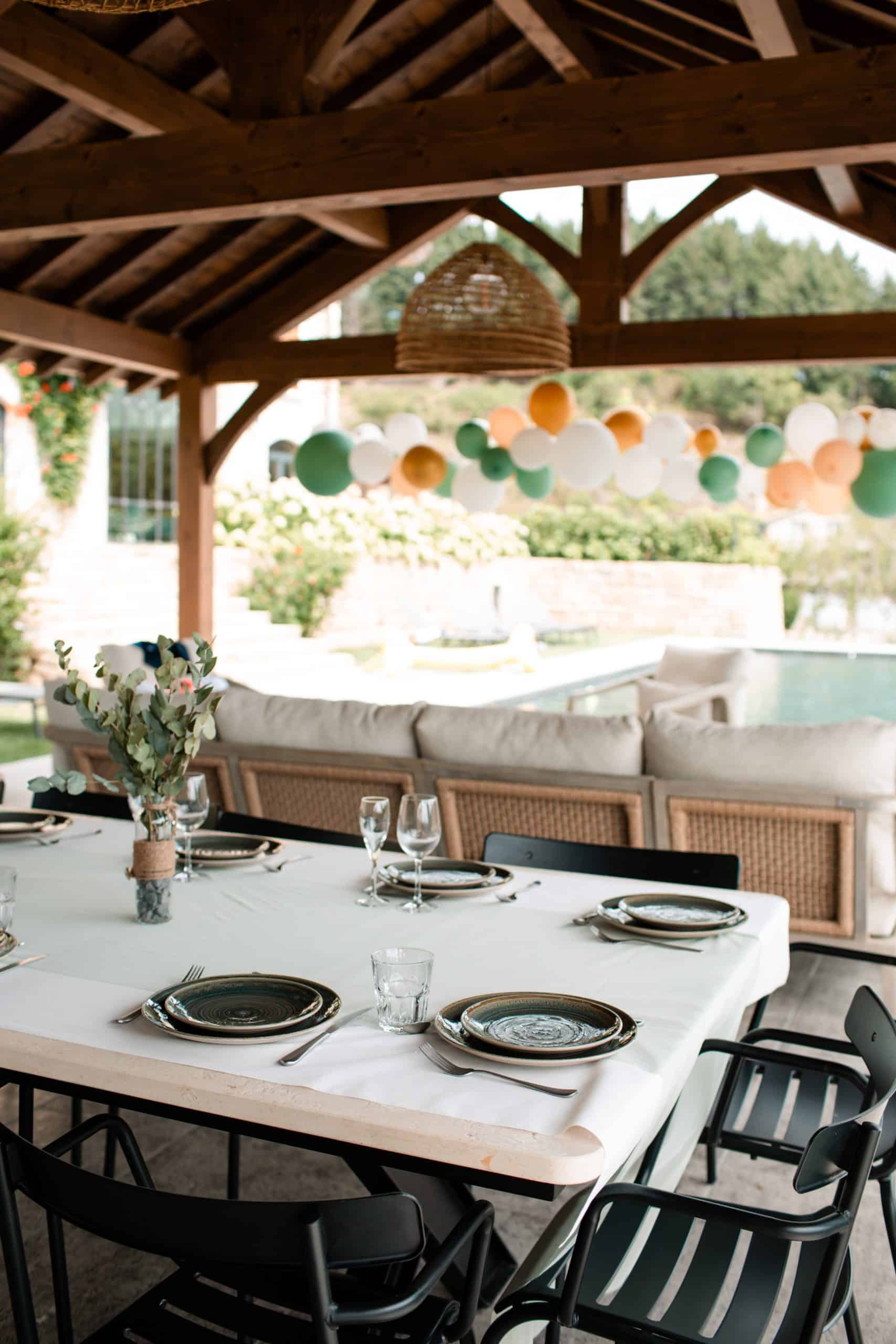 Une longue table dressée pour un repas sous une pergola en bois, décorée de ballons verts, oranges et blancs, avec une piscine en arrière-plan, dans une ambiance festive en extérieur.