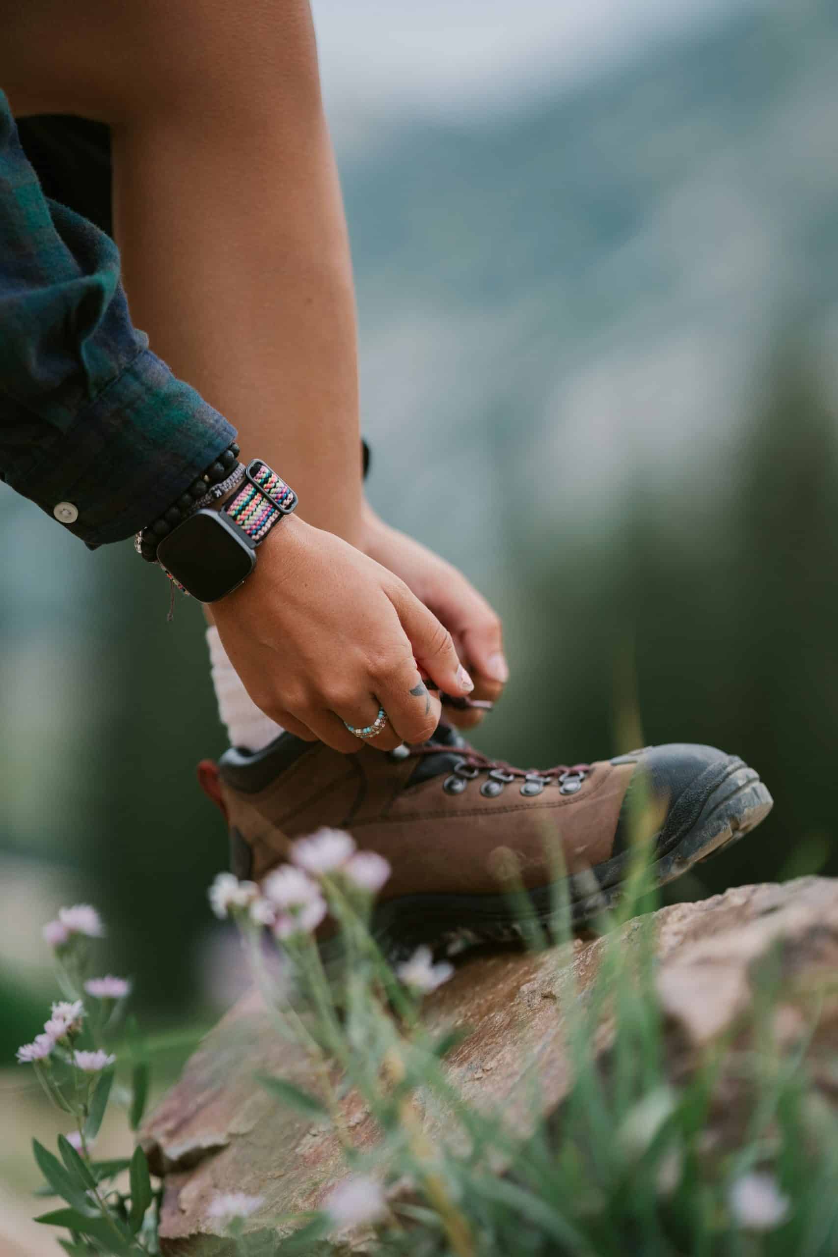 Une personne noue les lacets de sa botte de randonnée marron. Elle porte une chemise à carreaux, une montre connectée et des bracelets perlés, debout sur une roche avec des fleurs.
