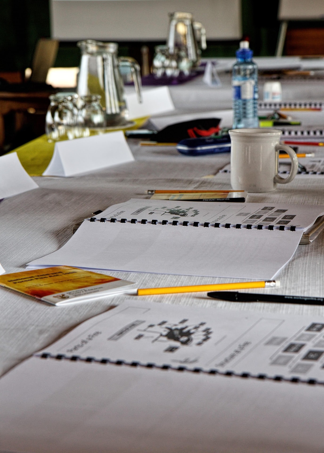 Plusieurs carnets à spirales ouverts, tasses, bouteille d'eau et fournitures d'écriture sont disposés sur une table claire, suggérant une conférence ou une réunion. Texte visible : Sur un crayon : "PREVENT WILDFIRE" Sur un stylo : "www.workingminds.org"