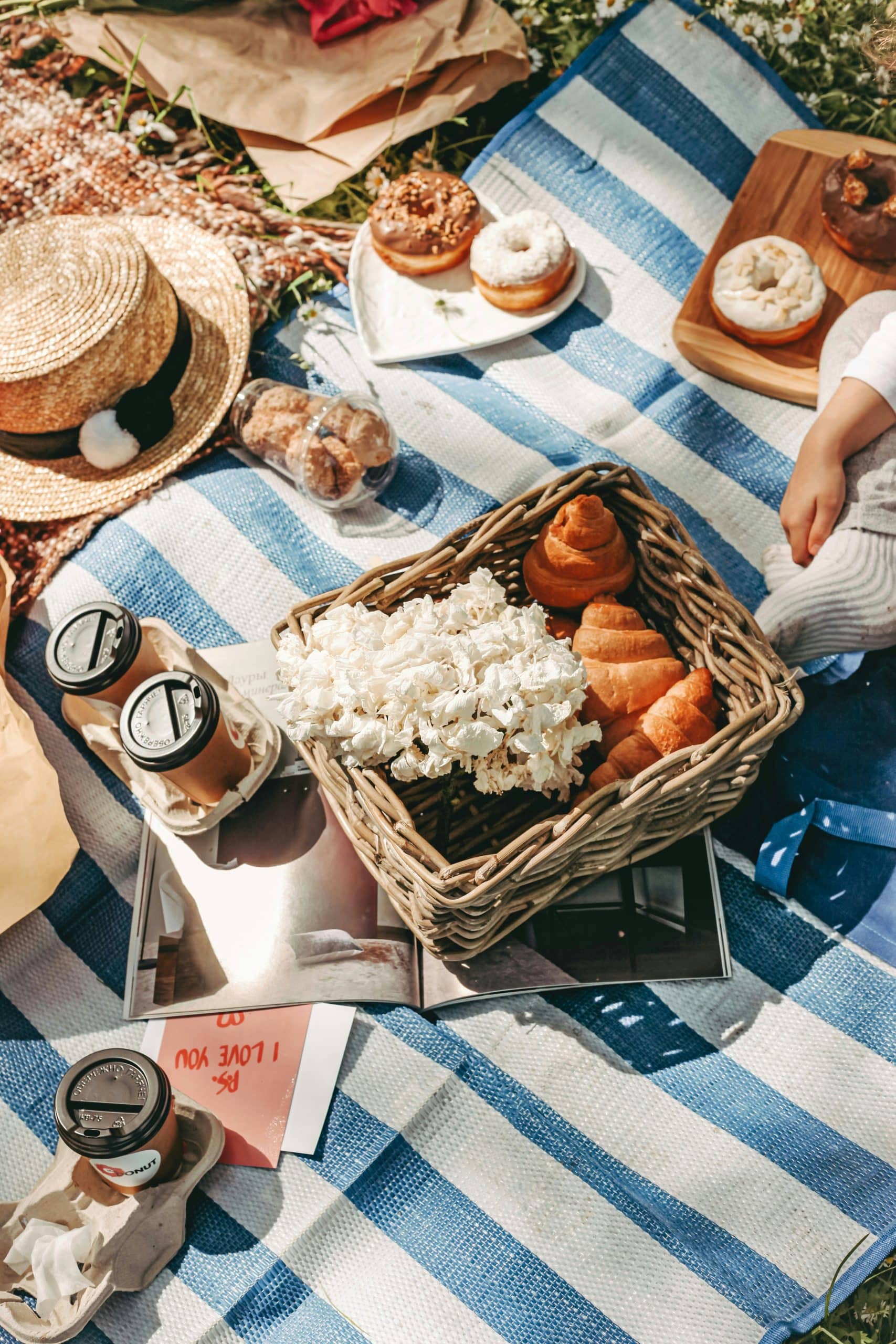 Pique-nique d'été avec donuts, croissants, café, fleurs et un chapeau disposés sur une couverture rayée bleue et blanche en extérieur, près d'une main d'enfant. Texte : « P.S. I LOVE YOU », « ОБЕРЕЖНО ГАРЯЧЕ! », « ДОНУТ ».