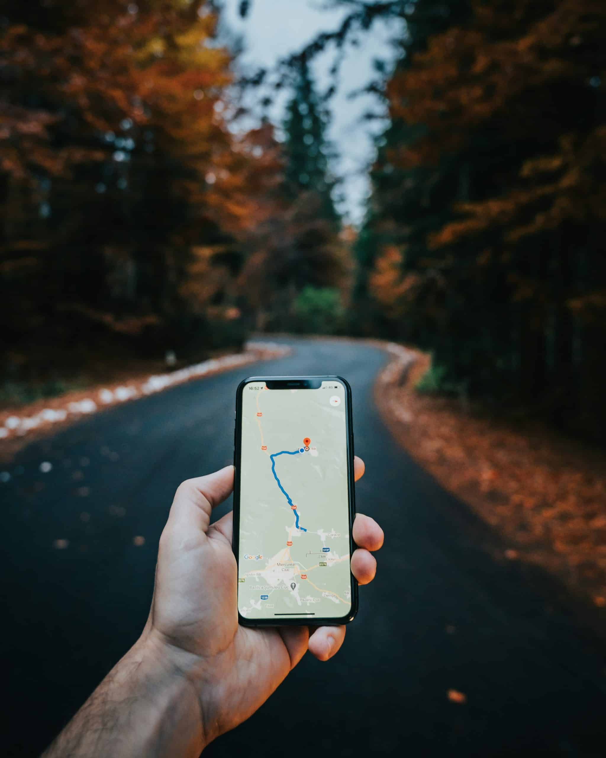 Une main tient un smartphone affichant un itinéraire GPS sur une route sinueuse bordée d'arbres aux couleurs automnales. Sur l'écran : 16:52, Wi-Fi, 4G, batterie, Google, Miercurea Ciuc, 13A, 123F, E578.