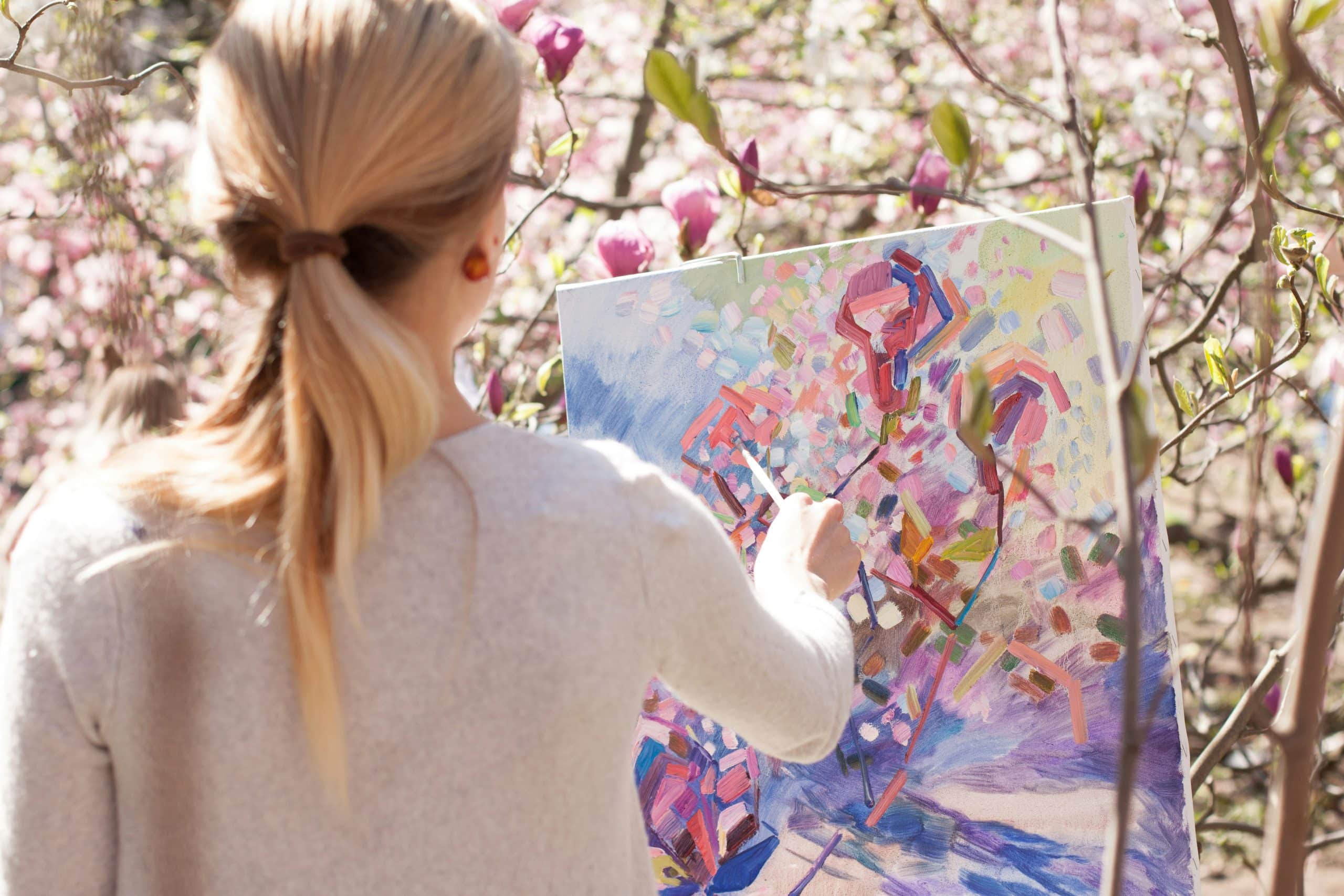 Une femme blonde, vue de dos, peint une toile abstraite et colorée avec un pinceau. Elle est dans un jardin fleuri de magnolias roses, sous le soleil.